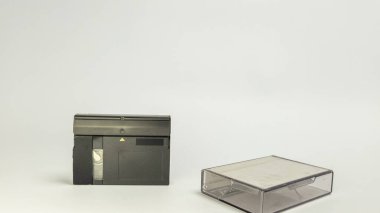 Eski bir MiniDV kaset kaydedicisinin ve onun temiz beyaz bir yüzeye yerleştirilmiş koruyucu çantasının yakın plan çekimi. Kayıt cihazı siyah ve dikdörtgen, kaseti yerleştirmek için bir yuvası var. Çanta şeffaf, izleyicinin görebilmesini sağlıyor.