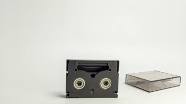 Beyaz arka planda koruyucu şeffaf plastik çantasıyla siyah bir Mini DV kasetin yakın çekim çekimi. Kaset dik konumda. Makara açılışlarıyla ön tarafını gösteriyor. Dava onun yanında duruyor. Görüntü biziz.