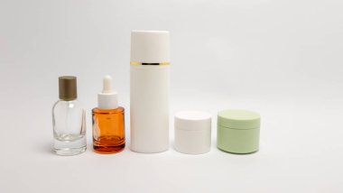 Temiz bir sprey şişesi, bir damlatıcı şişesi, uzun beyaz bir şişe ve sade beyaz arka planda düzenlenmiş iki küçük kavanoz içeren kozmetik şişe ve kavanoz koleksiyonu. Görüntü, cilt ürünleri için kullanılan konteynırların çeşitliliğini vurgular. Şey...