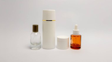 Bir sprey şişesi, altın çerçeveli uzun bir şişe, küçük bir kavanoz ve bir damla şişesi de dahil olmak üzere bir cilt ürünleri koleksiyonu el değmemiş beyaz bir yüzeye yerleştirilmiştir. Görüntü, temiz çizgiler ve minimalist çizgilerle aydınlık.