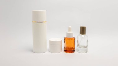 Beyaz arka planda sergilenen bir cilt bakım ürünleri koleksiyonu, altın bantlı uzun beyaz bir şişe, küçük beyaz bir kavanoz, kehribar sıvılı bir damla şişesi ve bir sprey şişesi yer alıyor. Kompozisyon temiz ve modern, temsil etmek için ideal. 
