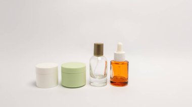 Temiz beyaz bir yüzeye yerleştirilmiş cilt bakım ürünleri koleksiyonu. Kompozisyonda iki krema kavanozu, saydam bir sprey şişesi ve bir kehribar cam damlası şişesi yer alıyor. Görüntü minimalist bir estetik taşıyor, ürünlerin basit el hareketlerini vurguluyor.