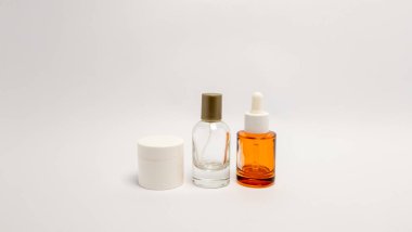 Sade beyaz arka planda üç kozmetik ürünü düzenlenir. Ürünler arasında beyaz bir kavanoz, kahverengi kapaklı berrak bir şişe ve damlatan turuncu bir cam şişe yer alıyor. Görüntü temiz ve basit, güzellik ve cilt pazarlaması için uygun..