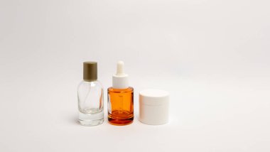 Minimalist bir canlı kompozisyonu, temiz beyaz arka planda üç cilt ürünü sergiliyor. Görüntüde açık bir cam sprey şişesi, canlı bir portakal damlası şişesi ve beyaz bir kavanoz yer alıyor. Kompozisyon, estetik çekiciliğe odaklanır.
