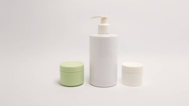 Bu resim, temiz beyaz bir arkaplana karşı düzenlenmiş bir pompa şişesi ve iki küçük kavanoz da dahil olmak üzere kozmetik ürünleri koleksiyonunu içeriyor. Kompozisyon, minimalist estetiği vurgulayarak ürünlerin tasarımı ve ambalajını vurguluyor. Işık.