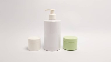 Pompalı beyaz bir losyon şişesi ve beyaz ve yeşil renkli iki küçük krema kavanozu olan bir stüdyo resmi. Ürünler, minimalist tasarımlarını ve cilt bakımında veya güzellik rutinlerinde potansiyel kullanımlarını vurgulayan temiz, beyaz bir yüzeye yerleştirilmiştir. İmaj