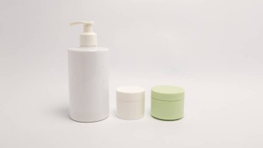 Üç kozmetik konteynırın yer aldığı bir stüdyo resmi: bir beyaz pompa şişesi ve iki küçük kavanoz, biri beyaz ve biri açık yeşil. Tüm konteynırlar düz beyaz arka planda. Işıklandırma yumuşak, minimalist tasarımı vurguluyor ve güzelliğe odaklanıyor.