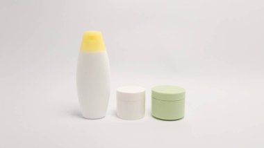 Bir minimalist, biri beyaz diğeri açık yeşil olmak üzere, sarı kapaklı beyaz bir şişe ve iki küçük yuvarlak kap içeren üç adet cilt bakım ürünü içerir. Ürünler düz beyaz bir arkaplan üzerine kuruludur. Görüntü basitliği vurgular