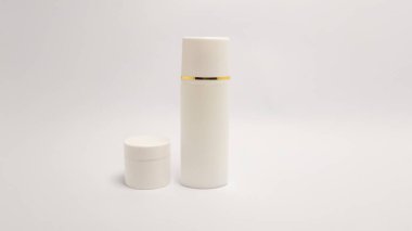 Bir stüdyo çekiminde düz beyaz arka plan üzerinde iki beyaz kozmetik kap bulunur. Uzun şişenin altın bir aksanı var ve görüntü profesyonelce aydınlatılmış. Temiz, minimalist estetik güzellik ürünü pazarlama ve cilt bakımı için idealdir.