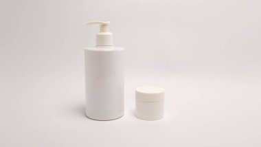Biri pompa şişesi, diğeri de kapaklı küçük bir kavanoz olan iki beyaz kozmetik kutunun minimalist stüdyo görüntüsü. Eşyalar beyaz bir zemine yerleştiriliyor, düz beyaz bir zemine karşı. Aydınlatma eşit ve kompozisyon basit, vurgula