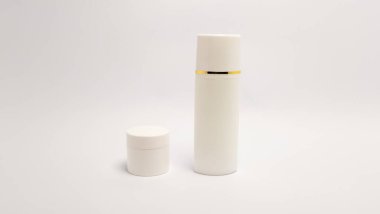 Beyaz arka planda iki beyaz kozmetik konteynırın yer aldığı minimalist bir stüdyo resmi. Ögeler uzun silindirik bir şişe, kapağı ve küçük bir kavanozu var. Işıklandırma yumuşak ve eşit, temiz çizgiler ve basit tasarım