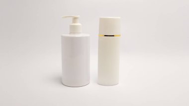 İki beyaz kozmetik şişesi düz beyaz arka planda yan yana duruyor. Bir şişe pompalı bir şeker şişesi. Diğer şişenin tepesinde altın bir şerit var. Şişeler temiz ve basit. Doğal güzelliğe odaklandığını gösteriyor.