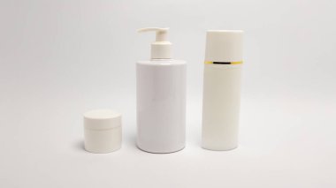 Üç beyaz kozmetik konteynırın yer aldığı bir stüdyo resmi: küçük bir kavanoz, bir pompa şişesi, ve üzerinde altın bir bant bulunan uzun bir şişe, hepsi düz beyaz bir fon üzerine kurulmuş. Basit düzenleme ve nötr tonlar, b için ideal, temiz ve modern bir estetik yaratır.