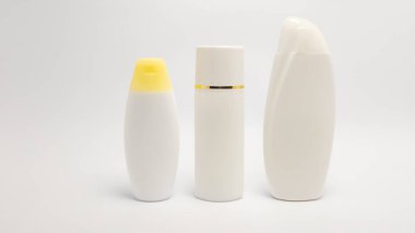 Üç beyaz kozmetik şişesi ardarda, temiz beyaz arka planda sıralanır. Şişeler şekil ve boyut olarak değişir ve birinin sarı bir kapağı vardır. Ortadaki şişenin boynunda altın bir şerit var. Görüntü minimalist bir estetiğe sahip, b için uygun.