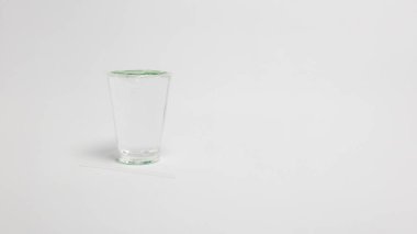 Açık, boş, yeşil kapaklı ve yanında şeffaf bir pipet bulunan bir bardak. Cam, tasarım veya metin yerleştirme için geniş negatif alan sağlayan temiz beyaz bir zemin üzerine yerleştirilmiştir. Görüntü iyi aydınlatılmış, aktarımı vurguluyor