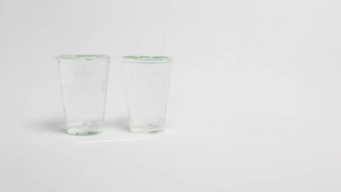 Bir stüdyo çekiminde iki temiz cam bardak ve el değmemiş beyaz bir fonda iki pipet bulunur. Bardaklar boş, basitliğe ve içecek potansiyeline odaklanmış. Aydınlatma parlak ve eşit, şeffaflığı vurguluyor