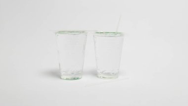 Bu stüdyo çekiminde içi su dolu iki şeffaf plastik bardak var. Bir bardak bir pipet yerleştirirken, bir pipet de yüzeyde duruyor. Bardaklar, temiz ve minimalist bir estetik oluşturan düz beyaz bir zemine yerleştirilmiş. İmaj