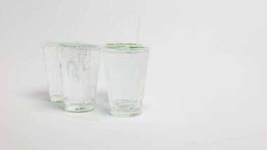 Beyaz bir yüzey üzerinde temiz sıvı, muhtemelen su ile dolu üç şeffaf plastik bardağın stüdyo çekimi. Bardakların kapakları var ve birinin içine pipet yerleştirilmiş. Bardakların önünde iki pipet daha var. Kompozisyon basit, t 'ye odaklı.