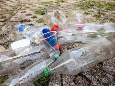 Çöpe atılmış plastik şişeler ve beton yüzeyde duran plastik bardaklar. Görüntü, plastik kirliliği ve çevre üzerindeki etkisini vurguluyor. Sahnede temiz plastik şişeler, çeşitli renkli kapaklar ve...