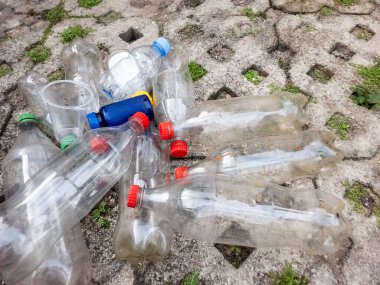 Yakın planda atılan plastik şişeler ve beton yüzeyde duran plastik bardaklar görülüyor. Şişeler çeşitli boyutlarda, bazıları kırmızı, mavi, yeşil ve berrak kapaklı. Sahne çevresel kirlilik ve atık yönetimi öneriyor