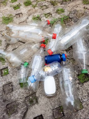 Yüksek açılı bir çekim, beton bir yüzeye saçılmış plastik şişe koleksiyonunu gösteriyor. Çeşitli boyutlarda ve şekillerde şişeler görülebilir, kırmızı, mavi ve yeşil kapaklı. Bazı şişeler boş, diğerleri ise orijinallerinin kalıntılarını taşıyor olabilir.