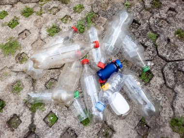 Tepeden çekilen bir resimde bir yığın plastik şişe ve beton bir yüzeye saçılmış bir bardak görülüyor. Şişeler kırmızı, yeşil ve mavi kapsüllerle boyut ve renk olarak farklılık gösterir ve bazıları kalıntı içerir. Sahne, plastik atık sorununu vurguluyor.