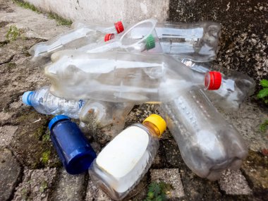 Çöpe atılmış plastik şişe ve konteynırlar çatlak bir kaldırımda yatıyor ve çevre kirliliği sorununu vurguluyor. Görüntü, su şişeleri ve şişe kapakları da dahil olmak üzere çeşitli plastik atık türlerini sergiliyor.