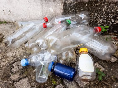 Çöpe atılmış plastik şişeler, bir fincan ve beton bir yüzeyi kirleten diğer konteynırlar. Görüntü, plastik atık ve kirliliğin çevresel sorununu vurguluyor. Şişelerin farklı renkte kapakları vardır. Bu fotoğraf bir kıskançlık hissi uyandırıyor.