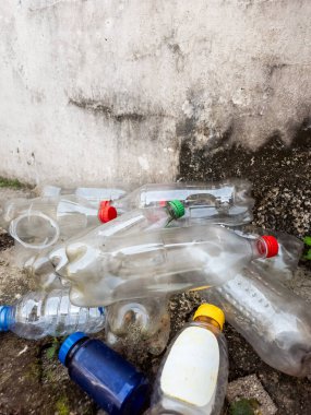 Yıpranmış beton bir duvara karşı yerde yatan bir yığın plastik şişenin yakın plan çekimi. Renkli kapaklı şeffaf şişeler ve mavi hap şişesi gelişigüzel dağıtılmış. Görüntü atık adam eksikliğini gösteriyor.