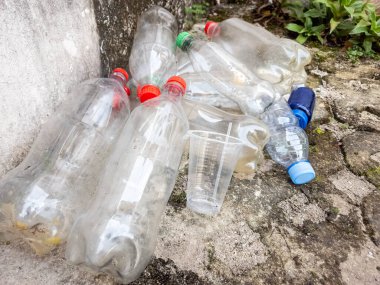 Yakın planda atılan plastik şişeler ve sert bir beton yüzeye saçılmış plastik bardaklar görülüyor. Şişeler çeşitli boyut ve renklerde, kırmızı, mavi ve yeşil kapaklı. Sahne, plastik kirliliği sorununu vurguluyor 