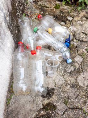Dökülen plastik şişeler ve plastik bardaklar desenli beton bir yüzey üzerinde duruyor. Görüntü, tek kullanımlık plastiklerle ilişkili atık ve kirliliği vurguluyor. Şişeler temiz ve boş, farklı renklerde kapakları var. Sahne gösteriyor ki