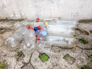 Çöpe atılmış plastik şişeler, kapaklar ve bir fincan yıpranmış beton bir yüzeye saçılmış. Görüntü, plastik atık ve çevre kirliliği sorununu vurguluyor. Kompozisyonda plastik atıkların beton bir duvara dayandığı görülüyor.
