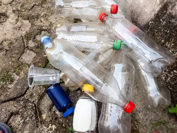 Yakın çekimde, kırık beton bir yüzeye saçılmış, atılmış plastik şişeler ve diğer plastik eşyalar görülüyor. Şişelerin farklı renkte kapakları var ve bazıları kısmen dolu. Görüntü çevre kirliliğini vurguluyor