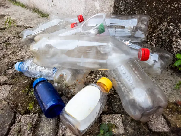 Çöpe atılmış plastik şişe ve konteynırlar çatlak bir kaldırımda yatıyor ve çevre kirliliği sorununu vurguluyor. Görüntü, su şişeleri ve şişe kapakları da dahil olmak üzere çeşitli plastik atık türlerini sergiliyor.