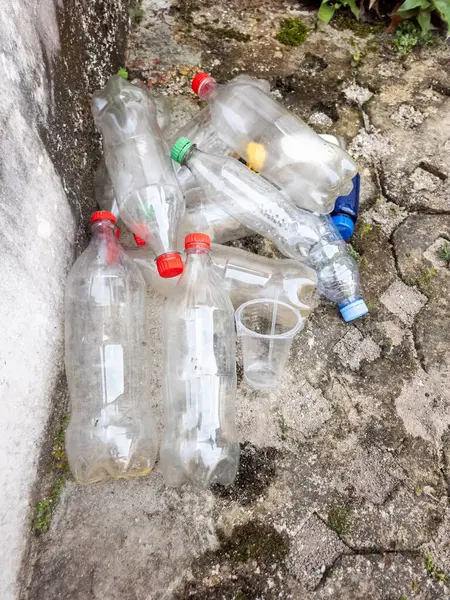 Dökülen plastik şişeler ve plastik bardaklar desenli beton bir yüzey üzerinde duruyor. Görüntü, tek kullanımlık plastiklerle ilişkili atık ve kirliliği vurguluyor. Şişeler temiz ve boş, farklı renklerde kapakları var. Sahne gösteriyor ki