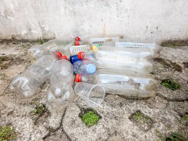 Çöpe atılmış plastik şişeler, kapaklar ve bir fincan yıpranmış beton bir yüzeye saçılmış. Görüntü, plastik atık ve çevre kirliliği sorununu vurguluyor. Kompozisyonda plastik atıkların beton bir duvara dayandığı görülüyor.