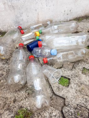 Çöpe atılmış plastik şişeler ve plastik bardaklar beton bir yüzey oluşturur. Görüntü, çevresel bir endişe ve kirlilik hissi veriyor. Kompozisyonda farklı renkte kapsüllere sahip çeşitli plastik kaplar yer alıyor. 