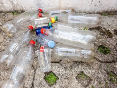 Yakından, yüksek açılı bir çekimde beton yüzeye saçılmış bir yığın plastik şişe, bardak ve diğer plastik atık görülüyor. Plastik farklı boyutlarda ve renklerde. Bu da çöp sorununun boyutunu gösteriyor. Resim vurguları