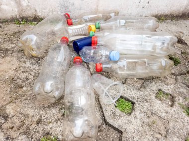 Bir yığın kullanılmış plastik şişe ve bir fincan çatlamış beton bir yüzeye saçılmış. Görüntü, plastik atık sorununu ve çevre üzerindeki etkisini vurguluyor. Çeşitli renkli şişe kapakları ve farklı şişe şekilleri var.