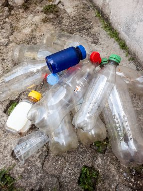 Yıpranmış beton bir yüzeye saçılmış plastik şişe ve konteynırların yakın plan çekimi. Resim, farklı renkli kapaklı berrak ve renkli şişeler de dahil olmak üzere çeşitli şekillerde ve boyutlarda plastik atık sergiliyor. Komşu.