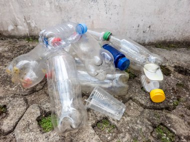 Çöpe atılmış plastik şişeler ve plastik bardaklar kaba beton bir yüzeye saçılmıştır. Görüntü, plastik kirliliği sorununu vurgulayarak, çevre sorumluluğuna açıkça saygısızlık gösteriyor. Kompozisyon yakın çekim.