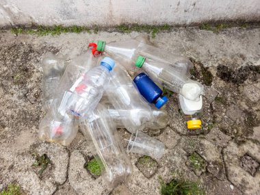 Çatlamış beton bir yüzeye saçılmış plastik şişeler ve plastik bardaklardan oluşan tepeden tırnağa bir görüntü. Açık plastik şişelerin farklı renkte kapakları var. Bu da farklı içecekleri gösteriyor. Görüntü, plastik atık ve çevre sorununu vurguluyor