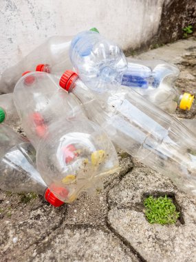 Beton bir yüzeye saçılmış kullanılmış plastik şişe koleksiyonu, plastik atık ve çevre kirliliği sorununu vurguluyor. Görüntüde renkli kapaklı çeşitli berrak şişeler ve Crac 'ta büyüyen küçük bir parça yeşillik yer alıyor.