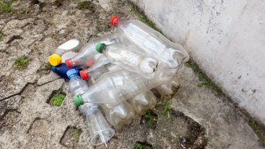 Çatlamış beton bir yüzey üzerinde, beton bir duvarın yanında duran boş ve temiz plastik şişelerin yakın çekim görüntüleri. Şişelerin çeşitli renkli kapakları vardır. Görüntü, biraz yeşil vejetaryenle plastik atık ve çevre kirliliği sorununu vurguluyor