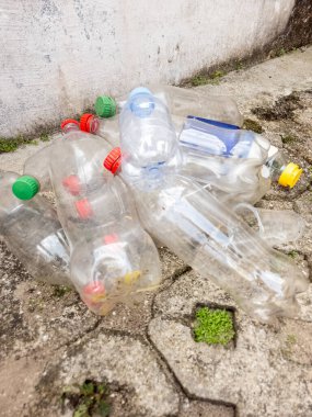 Çöpe atılmış plastik şişelerden oluşan bir kolleksiyon beton bir yüzeye saçılmış olarak görülür. Şişelerin çeşitli renkli kapakları var ve bakımsız durumdalar. Bu sahne plastik kirliliği ve çevresel atık sorununu vurguluyor.