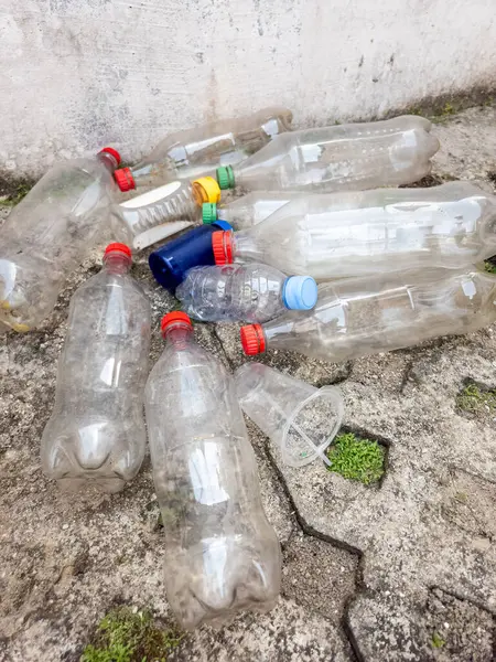 Çöpe atılmış plastik şişeler ve plastik bardaklar beton bir yüzey oluşturur. Görüntü, çevresel bir endişe ve kirlilik hissi veriyor. Kompozisyonda farklı renkte kapsüllere sahip çeşitli plastik kaplar yer alıyor. 