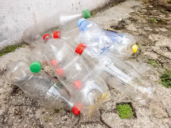 Kaba beton bir yüzeye atılmış renkli kapaklı temiz plastik şişeler yığını. Görüntü, çevre kirliliği ve atık yönetimi sorunlarını vurguluyor. Kompozisyon, plastiğin hacmini ve çevre üzerindeki etkisini vurgular.