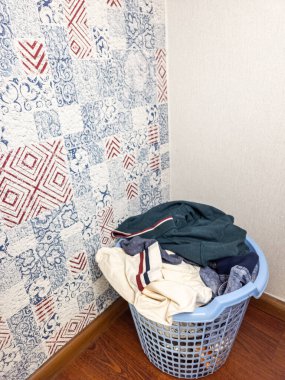 Elbiselerle dolu açık mavi bir çamaşır sepeti odanın köşesinde duruyor. Odada mavi ve kırmızı geometrik tasarımlı desenli duvar kağıdı ve ahşap zemin bulunuyor. Sepet çeşitli giysiler içeriyor, sıradan bir yaşam tarzı ve... 