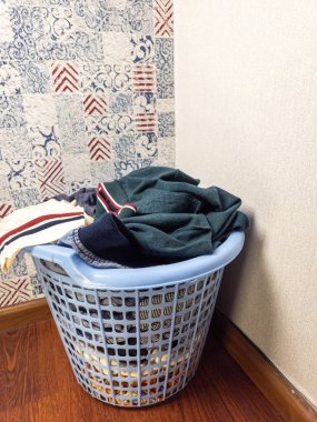 Elbiselerle dolu açık mavi bir çamaşır sepeti odanın köşesinde duruyor. Sepet ahşap bir zemine yerleştirilmiş ve duvarlar desenli bir duvar kağıdıyla boyanmış. Giysiler renklerin karışımı, ve sahne bir arifenin habercisi.