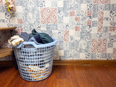 Temiz giysilerle dolu açık mavi bir çamaşır sepeti ahşap bir zeminde desenli bir duvar kağıdı arka planında duruyor. Sepet gömlek, pantolon ve diğer giysiler gibi eşyalarla dolup taşıyor. Görüntü, bir aile sahnesini yakalıyor.